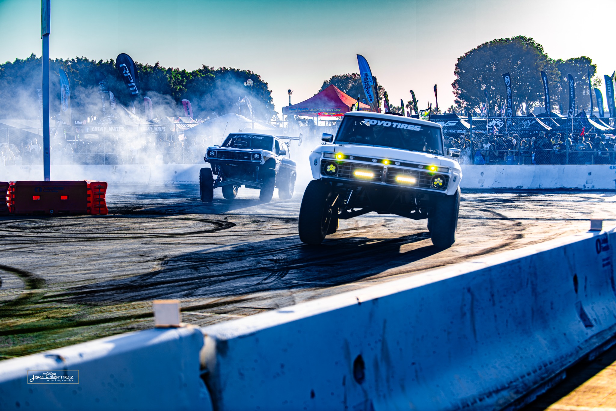 Two white off-road trucks cross the marked arena surface with smoke and spectators behind them