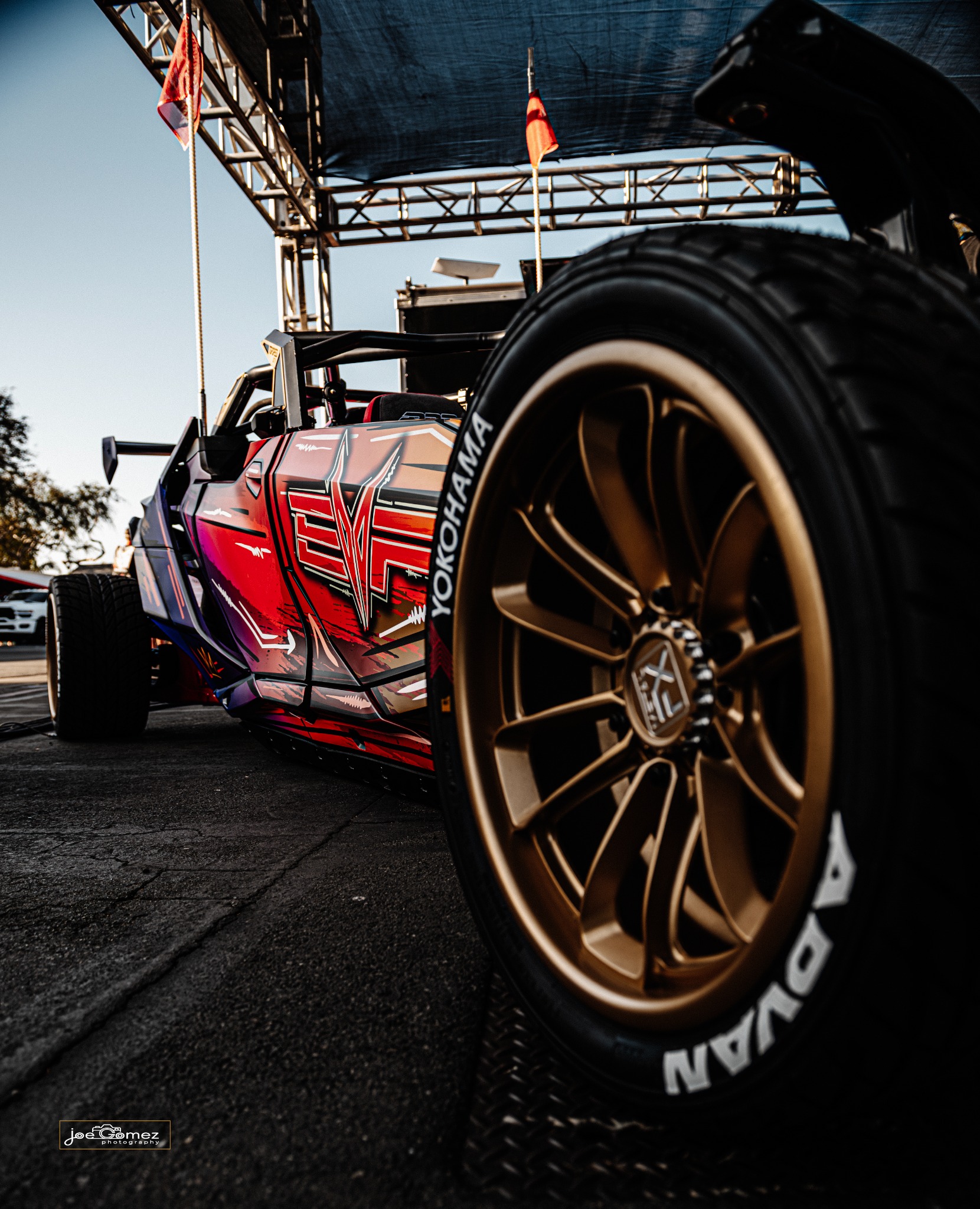 A bronze wheel fills the foreground beside a red-and-black off-road vehicle under a display canopy